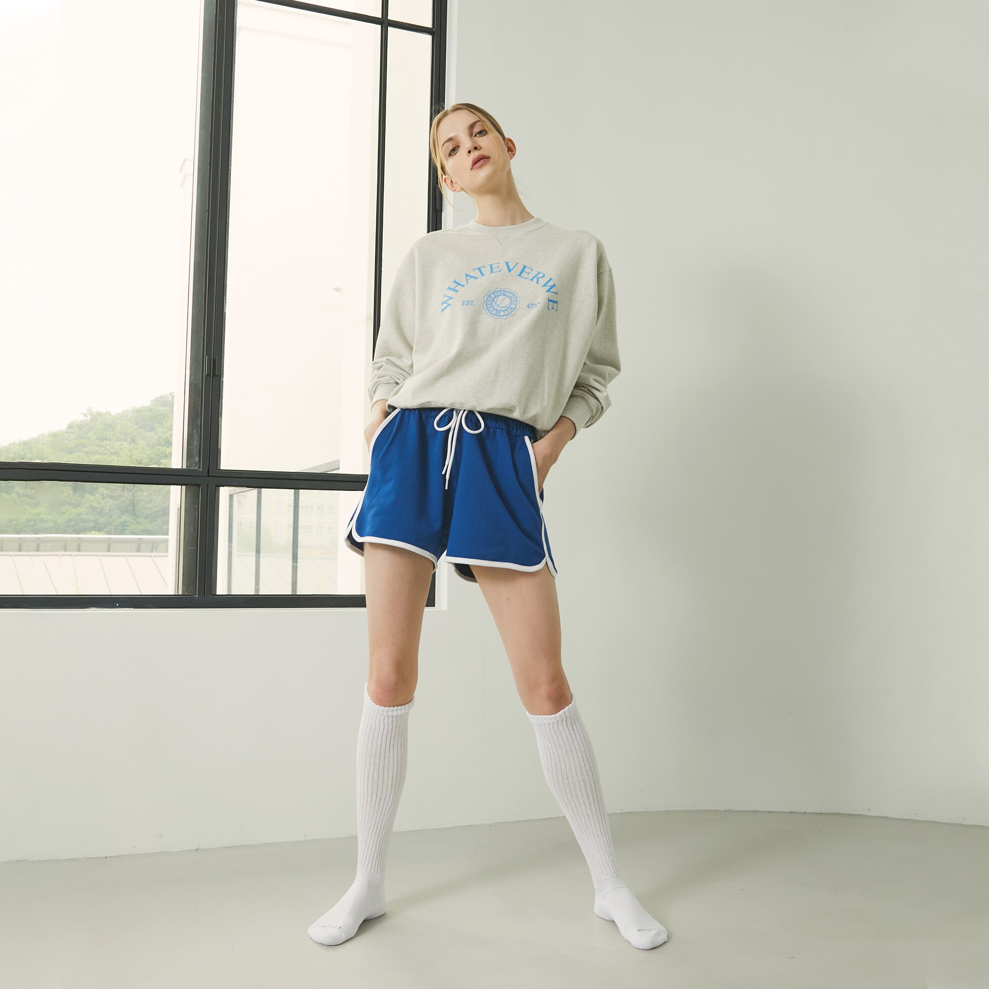 A woman standing in front of a window wearing a sweatshirt and shorts.