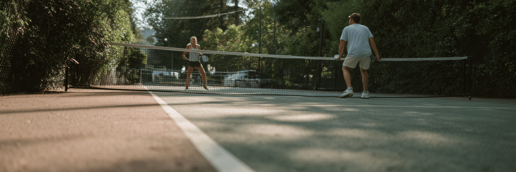 A man and woman stand on opposite sides of a net on an outdoor pickleball court surrounded by lush greenery.