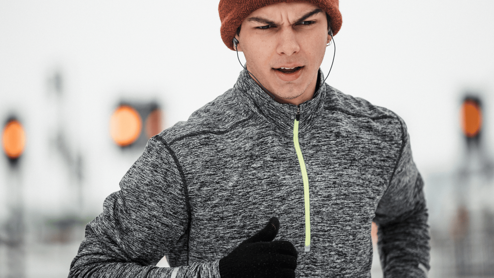 Man running outdoors in cold weather, wearing a gray athletic jacket, orange beanie, gloves, and earphones.