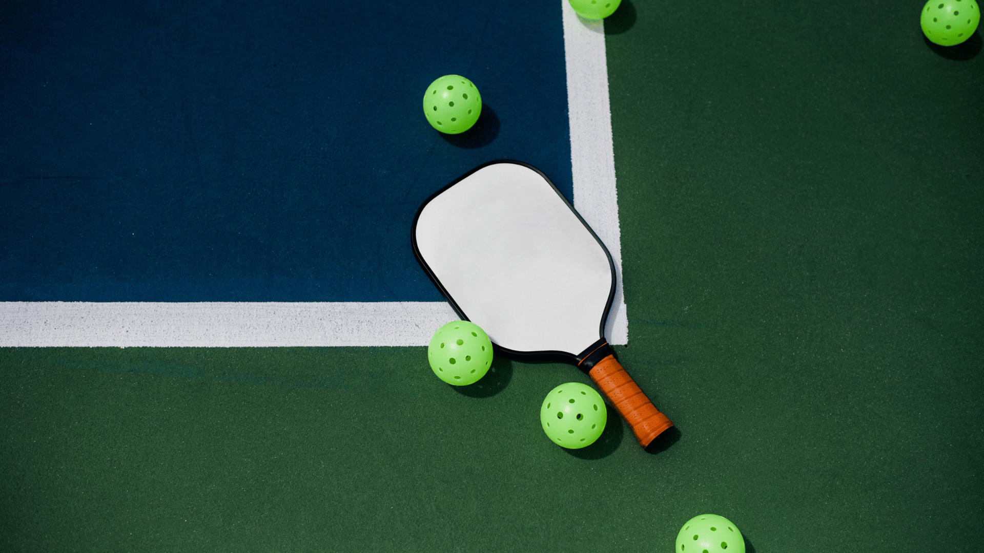 Pickleball paddle with an orange grip and neon green balls scattered on a blue and green court.