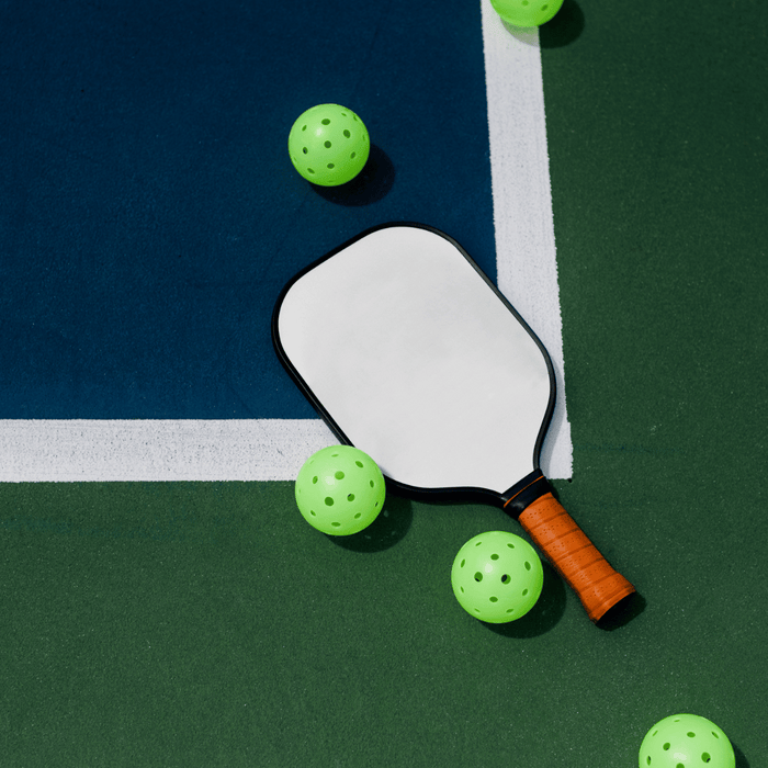 Pickleball paddle with an orange grip and neon green balls scattered on a blue and green court.