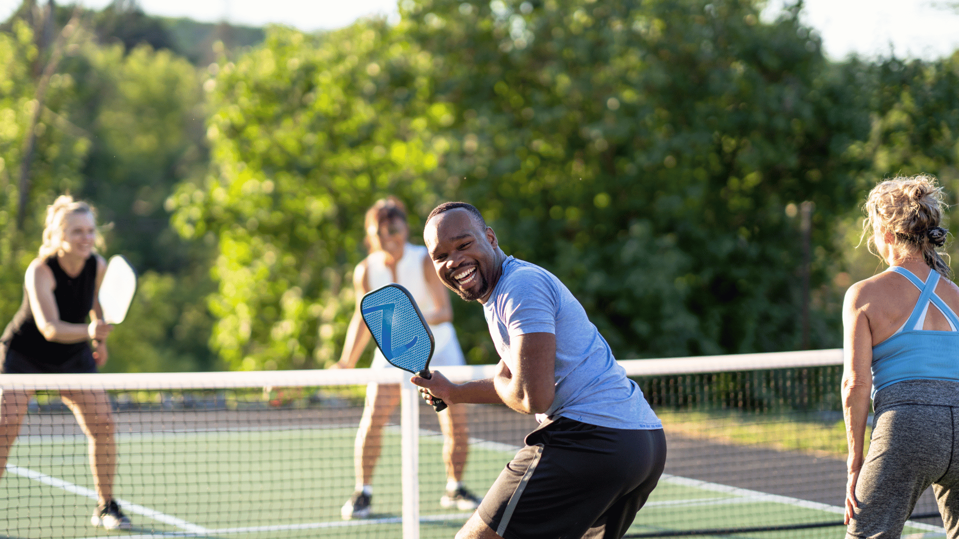 Group of four people playing pickleball outdoors on a sunny day, with one man smiling while holding a blue paddle near the net.