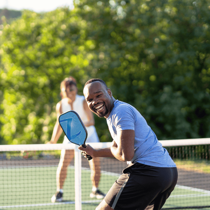 Group of four people playing pickleball outdoors on a sunny day, with one man smiling while holding a blue paddle near the net.