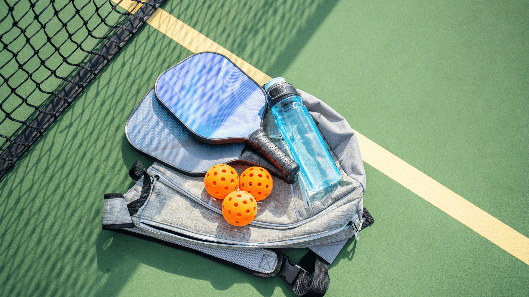 Pickleball gear including a blue paddle, three orange balls, a water bottle, and a backpack placed on a green court next to the net.