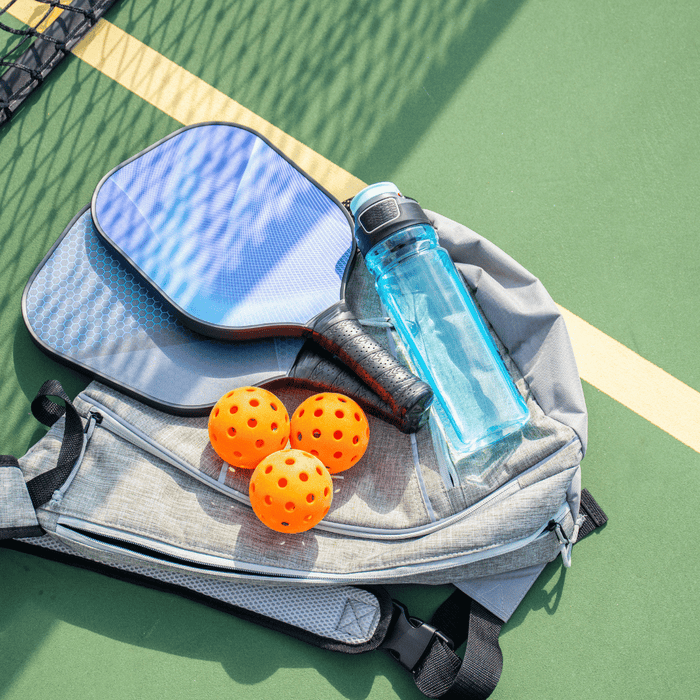 Pickleball gear including a blue paddle, three orange balls, a water bottle, and a backpack placed on a green court next to the net.