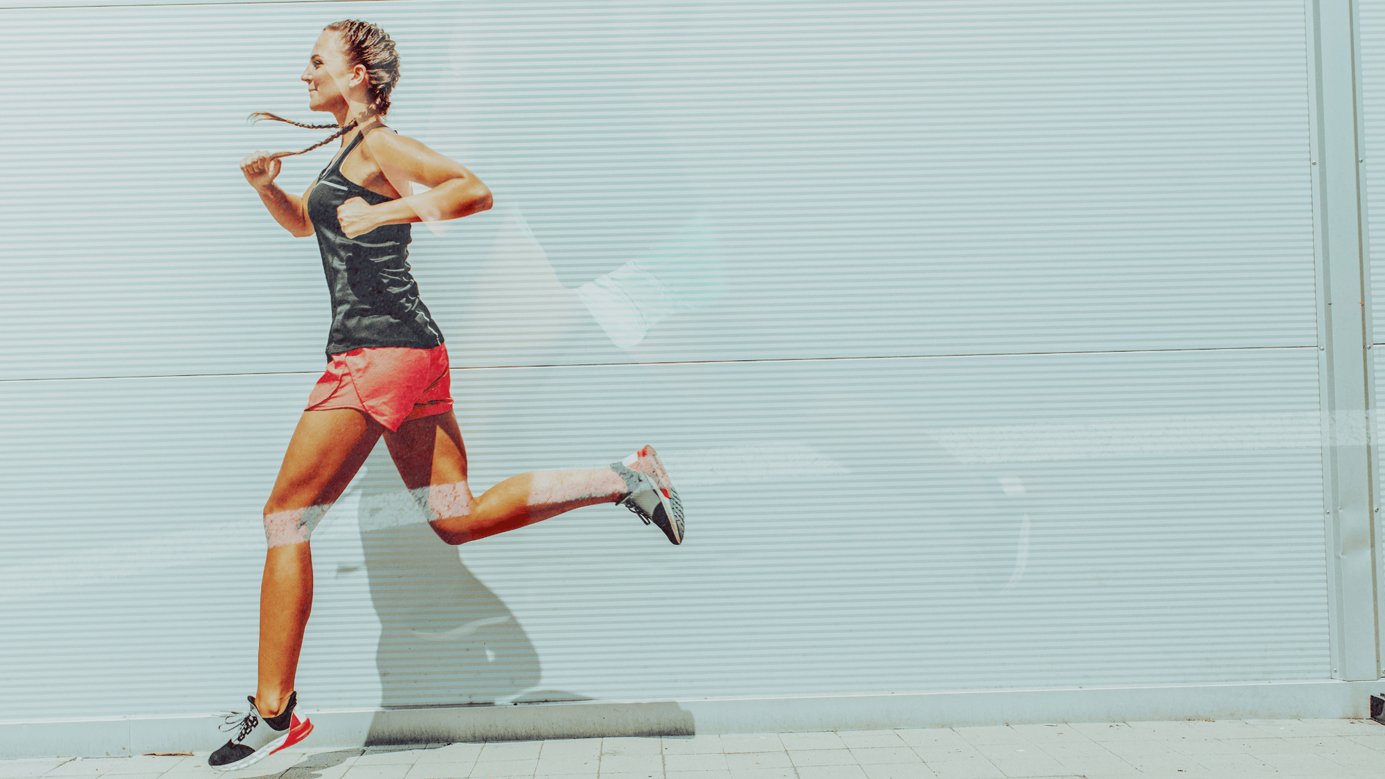 Woman in athletic gear running outdoors against a light corrugated wall, wearing a black tank top, red shorts, and running shoes.