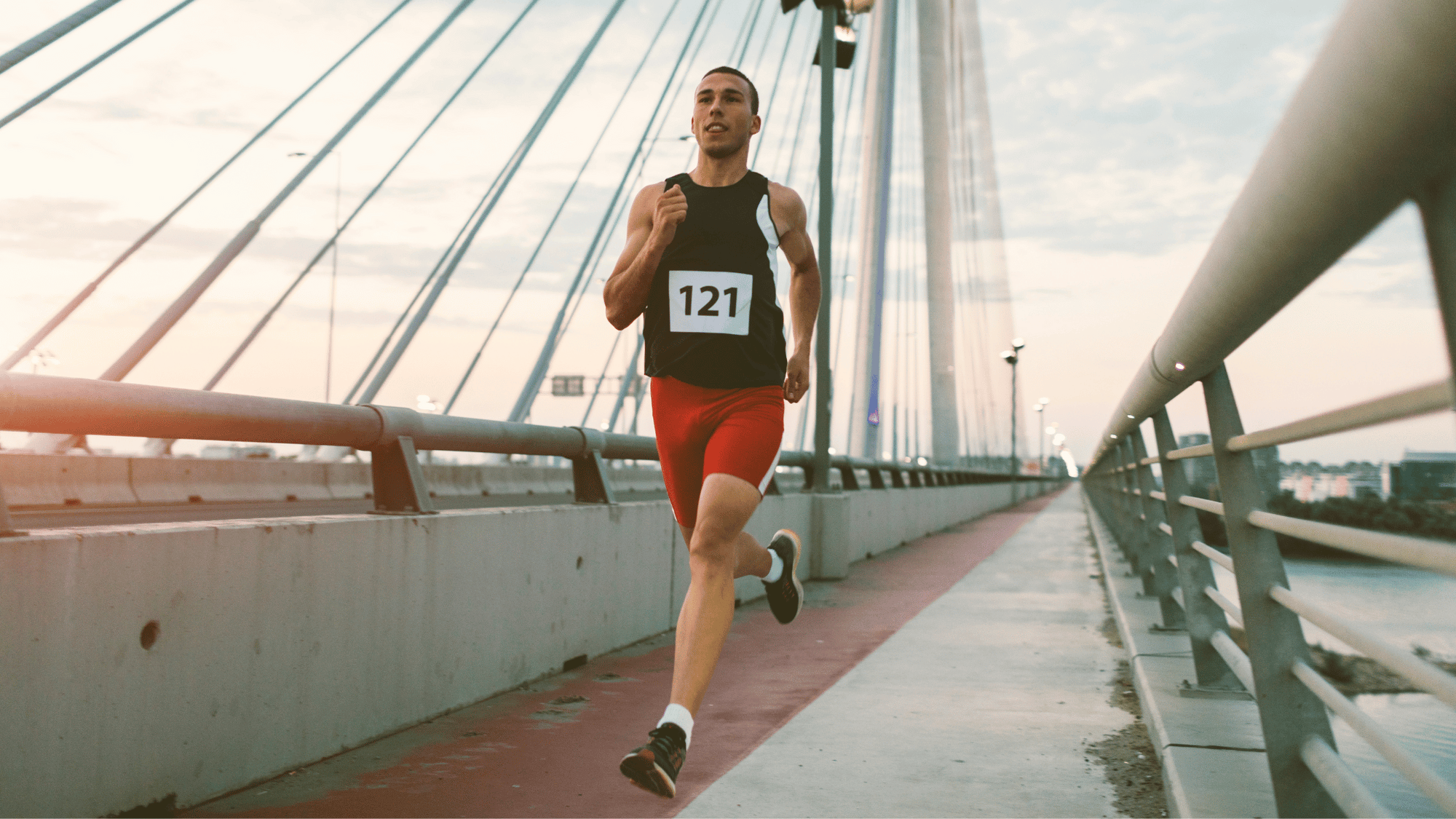 Runner in red shorts and tank top, number 121, sprinting on a cable-stayed bridge at sunset, focused and determined.