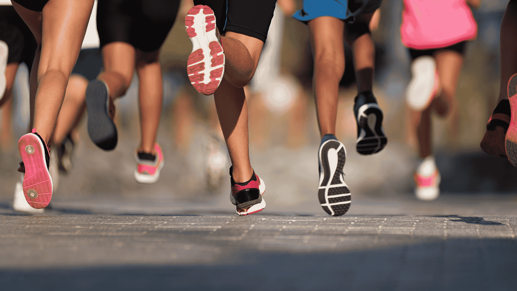 Group of runners in colorful shoes mid-stride, viewed from behind, showing dynamic movement on a sunny pavement.