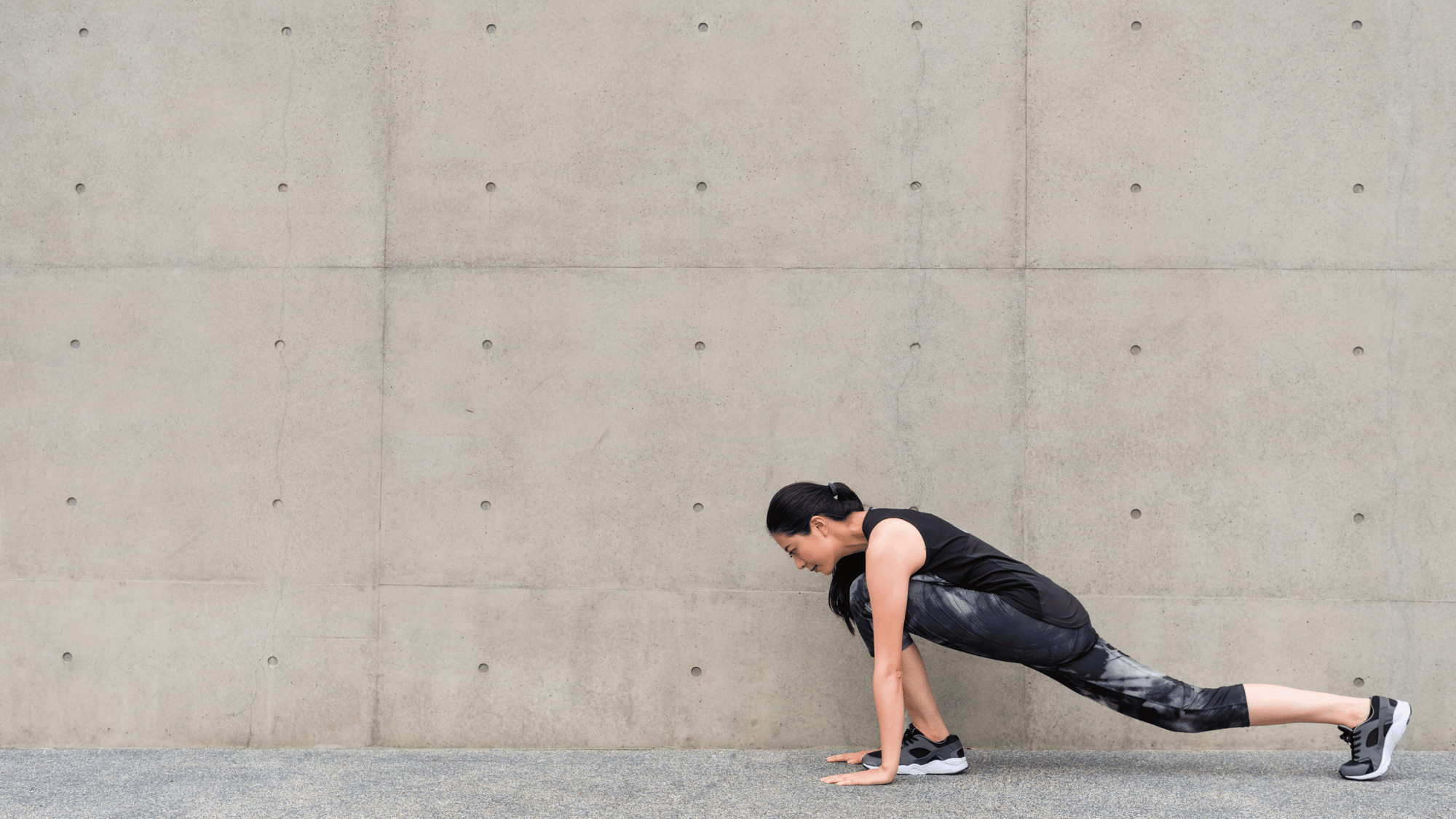 Woman in athletic wear performing a low lunge stretch against a textured concrete wall.